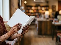 Woman with henna-decorated hands reading open book in modern bookstore or café setting with warm ambient lighting and blurred background.