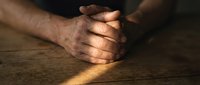 Adult male with medium brown skin tone, hands clasped together on wooden surface in prayer or meditation pose, warm natural lighting.