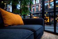 Modern navy blue sofa with orange accent pillow in contemporary urban setting, evening city lights and building reflections visible through large windows.