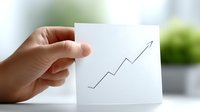 Hand holding white paper card with upward trending arrow graph showing business growth and success in bright office environment.
