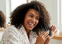 Happy African American woman with curly hair brushing her natural hair in bathroom mirror, smiling while doing morning hair care routine.