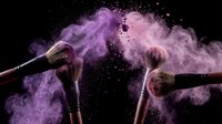 Makeup brushes with purple and pink powder explosion on black background creating dramatic cosmetic dust cloud effect for beauty industry.