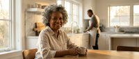Happy mature African American woman sitting at kitchen counter smiling warmly while man works in background during sunny morning at home.