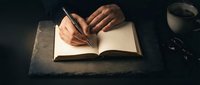 Young adult male with medium skin tone writing in open notebook with fountain pen at dark desk, coffee cup nearby, overhead view for journaling, blogging, education, and creative writing concepts.