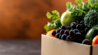 Fresh organic produce in cardboard box with grapes, apples, broccoli, cucumber and leafy greens against rustic brown background, selective focus on fruits and vegetables.