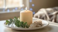Cozy home interior with burning candle, eucalyptus branch and decorative stones on white plate creating peaceful spa-like atmosphere for relaxation.