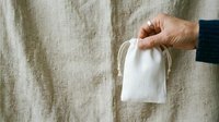 Adult hand holding white cotton drawstring pouch against natural linen fabric background. Suitable for eco-friendly product packaging, sustainable lifestyle content, and organic goods marketing.