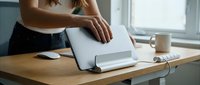 Woman with medium skin tone working at wooden desk, adjusting laptop stand for ergonomic posture. Modern home office setup with natural light, coffee cup, and wireless mouse for productivity and remote work.