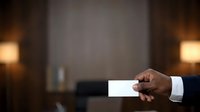 Male professional with dark skin tone in formal business attire holding blank white business card in office setting with warm ambient lighting and blurred background for corporate branding.