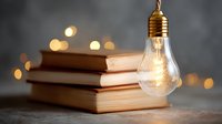 Glowing light bulb hanging above stack of vintage books with soft bokeh background creates atmospheric mood for education and inspiration concepts.