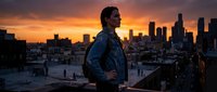 Young professional man standing on urban rooftop overlooking city skyline at golden hour sunset with dramatic orange sky and downtown buildings.