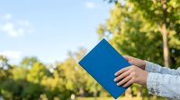 Person holding blue notebook outdoors with green trees and sky background, suitable for education, business, and lifestyle marketing materials.