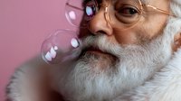 Senior Caucasian man with white beard and round glasses playfully blowing soap bubbles against pink background, expressing joy and childlike wonder in lifestyle portrait.