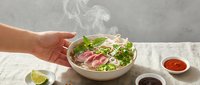 Young adult hand holding steaming bowl of Asian noodle soup with fresh greens and sliced meat, ready-to-eat meal preparation for food blogs and culinary content.