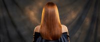 Young woman with long straight red hair viewed from behind wearing black off-shoulder top against dark dramatic background for beauty and fashion concepts.