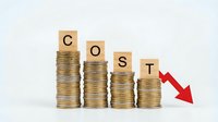 Business cost reduction concept with wooden letter blocks spelling COST on decreasing stacks of golden coins and red downward arrow showing financial savings.