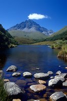 Serene mountain lake with rocky shoreline and majestic peak rising against vibrant blue sky, reflecting in calm waters surrounded by natural grassland landscape.