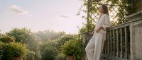 Young woman in flowing white dress standing on stone balcony with ornate railings surrounded by lush garden foliage during golden hour sunset light.