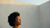 Young woman with dark curly hair and medium skin tone in profile, gazing upward against neutral beige and gray wall background during golden hour lighting.