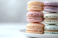 Colorful French macarons stacked on white plate against light background. Pastel pink, purple, peach and mint green cookies with cream filling create delicate dessert composition.