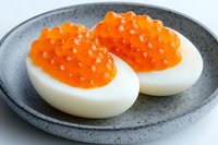 Halved boiled egg topped with bright orange salmon caviar served on gray ceramic plate, close up view of gourmet appetizer against light background.