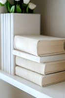 Stack of white hardcover books on shelf with minimalist aesthetic and soft natural lighting, green plant in background creates serene home library atmosphere.