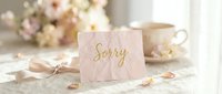 Soft pink sorry card with gold script lettering on elegant table with flowers and tea cup, apology and forgiveness concept.