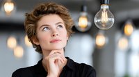Young professional woman thinking with hand on chin looking up at light bulb, creative inspiration and innovation concept in modern office environment.