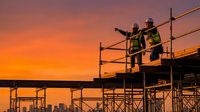 Construction workers on scaffolding at sunset discussing project plans with dramatic orange sky backdrop for industrial and building development concepts.