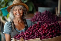 Mature female farmer in straw hat and overalls smiling while holding wooden crate full of freshly harvested purple grapes at local market or vineyard.