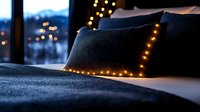 Cozy bed with glowing string lights on dark pillows against evening window view, creating romantic ambient atmosphere in bedroom interior.