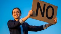Young woman with dark hair and light skin tone, wearing dark blazer, holding cardboard sign with NO text against blue sky background, expressing rejection or disagreement.