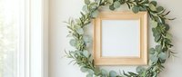 Wooden square frame surrounded by fresh eucalyptus and olive branches wreath on white wall with natural light, suitable for home decor and interior design projects.