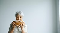 Senior woman with gray hair and light skin tone, wearing white shirt, holding and eating fresh baked bread loaf against white background for food and lifestyle content.