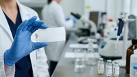 Healthcare professional in lab coat and blue gloves holding blank white card in modern laboratory setting with scientific equipment and bottles in background.