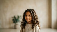 Young girl with long curly dark hair and medium skin tone wearing a cream ribbed sweater, smiling at camera in bright indoor studio setting with neutral background.