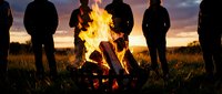 Group of friends gathered around crackling campfire at sunset in open field creating warm social atmosphere for outdoor adventure and bonding experiences.