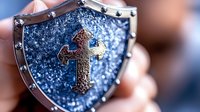 Medieval shield with ornate golden cross on sparkling blue background, metallic frame with rivets, close-up macro photography showing intricate details and texture.