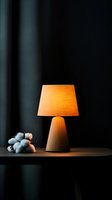 Wooden table lamp with orange fabric shade glowing warmly against dark background, accompanied by cotton plant stems creating cozy home atmosphere.