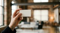 Hand holding blank white business card in modern office space with blurred interior background, suitable for branding and corporate identity mockups.