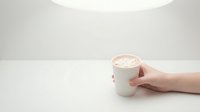Female hand holding white ceramic mug with frothy cappuccino on clean minimalist table surface with soft natural lighting for cafe branding materials.