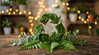 Fresh green fern leaves and eucalyptus branches arranged in rustic wreath on wooden table with warm bokeh lights creating cozy natural home decor atmosphere.