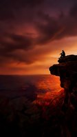 Silhouetted male figure sitting on rocky cliff edge overlooking vast canyon landscape during dramatic golden sunset with stormy clouds. Perfect for adventure, inspiration, and contemplation themes.