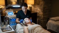 Professional skincare treatment session with female client receiving facial therapy from skilled aesthetician in modern spa setting with advanced equipment.