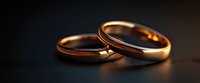Two elegant gold wedding bands with subtle diamond accent lying together on dark reflective surface, illuminated by warm dramatic lighting. Symbolizes marriage commitment.