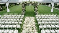 Elegant outdoor wedding ceremony setup with floral arch, white chairs, and petal-lined aisle under a transparent tent on lush green lawn.