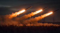 Multiple rockets launching at dusk with bright orange exhaust trails against dark dramatic sky, silhouetted grass in foreground creates striking military scene.