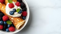 Fresh crepes filled with whipped cream, garnished with mixed berries and mint leaves on white plate, selective focus with copy space on marble background.