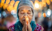 Elderly Asian woman with closed eyes praying, wearing traditional headscarf against blurred golden bokeh background, expressing deep spiritual devotion and mindfulness.
