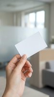 Hand holding blank white business card mockup in modern office interior with natural lighting for branding and corporate identity design projects.